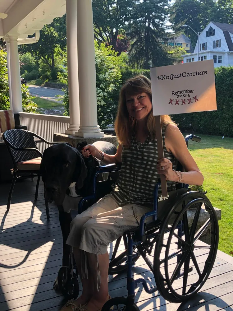 A woman in a wheelchair holding up a sign that reads "#NotJustCarriers" above the Remember The Girls logo.