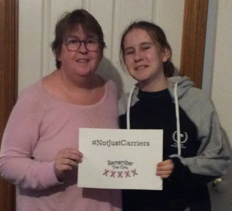 Two community members holding up a sign that reads "#NotJustCarriers" above the Remember The Girls logo.