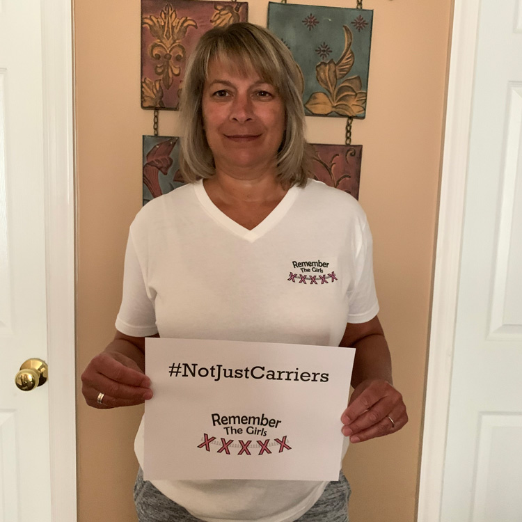 A community member in a Remember The Girls shirt holding up a sign that reads "#NotJustCarriers" above the Remember The Girls logo.