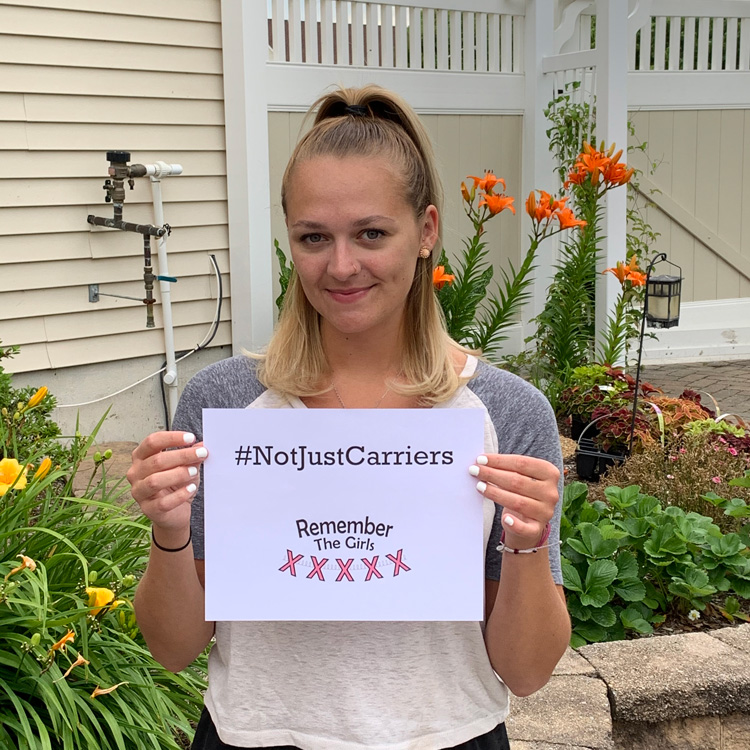 A woman in front of a garden holding up a sign that reads "#NotJustCarriers" above the Remember The Girls logo.
