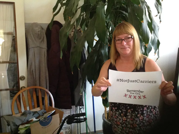 A community member in her house holding up a sign that reads "#NotJustCarriers" above the Remember The Girls logo.
