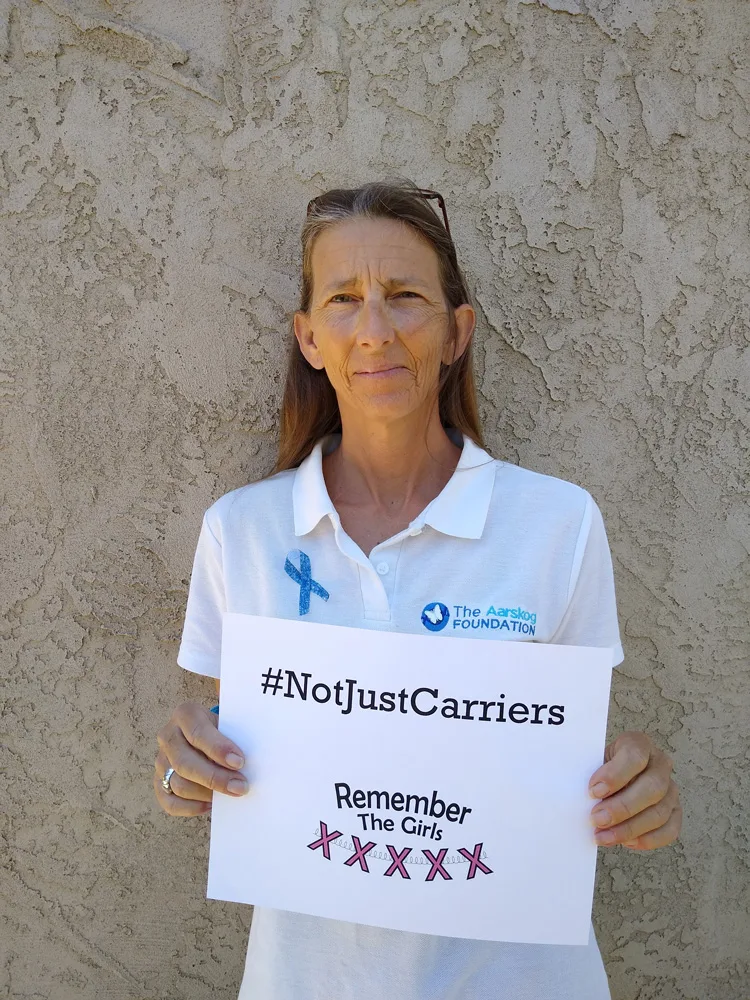 A woman in front of a wall wearing a shirt with a logo for The Aarskog Foundation holding up a sign that reads "#NotJustCarriers" above the Remember The Girls logo.