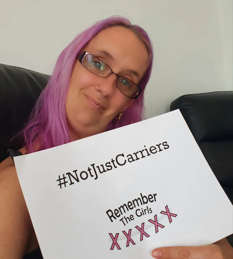A community member with glasses holding up a sign that reads "#NotJustCarriers" above the Remember The Girls logo.