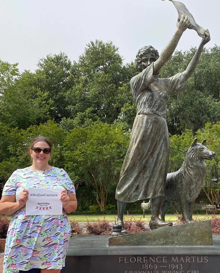A woman in front of a statue holding up a sign that reads "#NotJustCarriers" above the Remember The Girls logo.
