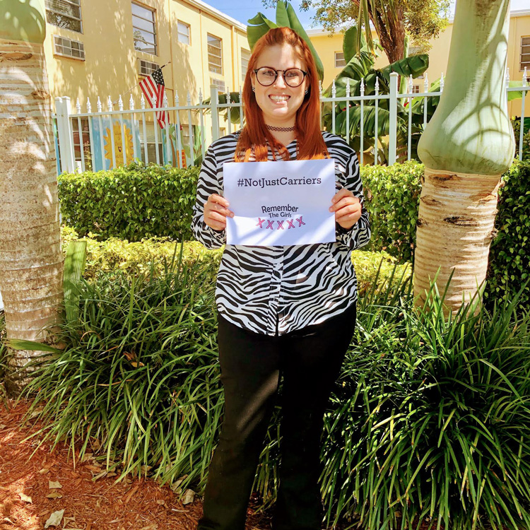 A community member in a zebra print shirt holding up a sign that reads "#NotJustCarriers" above the Remember The Girls logo.