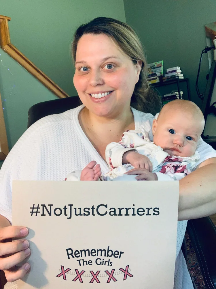 A community member holding her baby and a sign that reads "#NotJustCarriers" with a Remember The Girls logo.