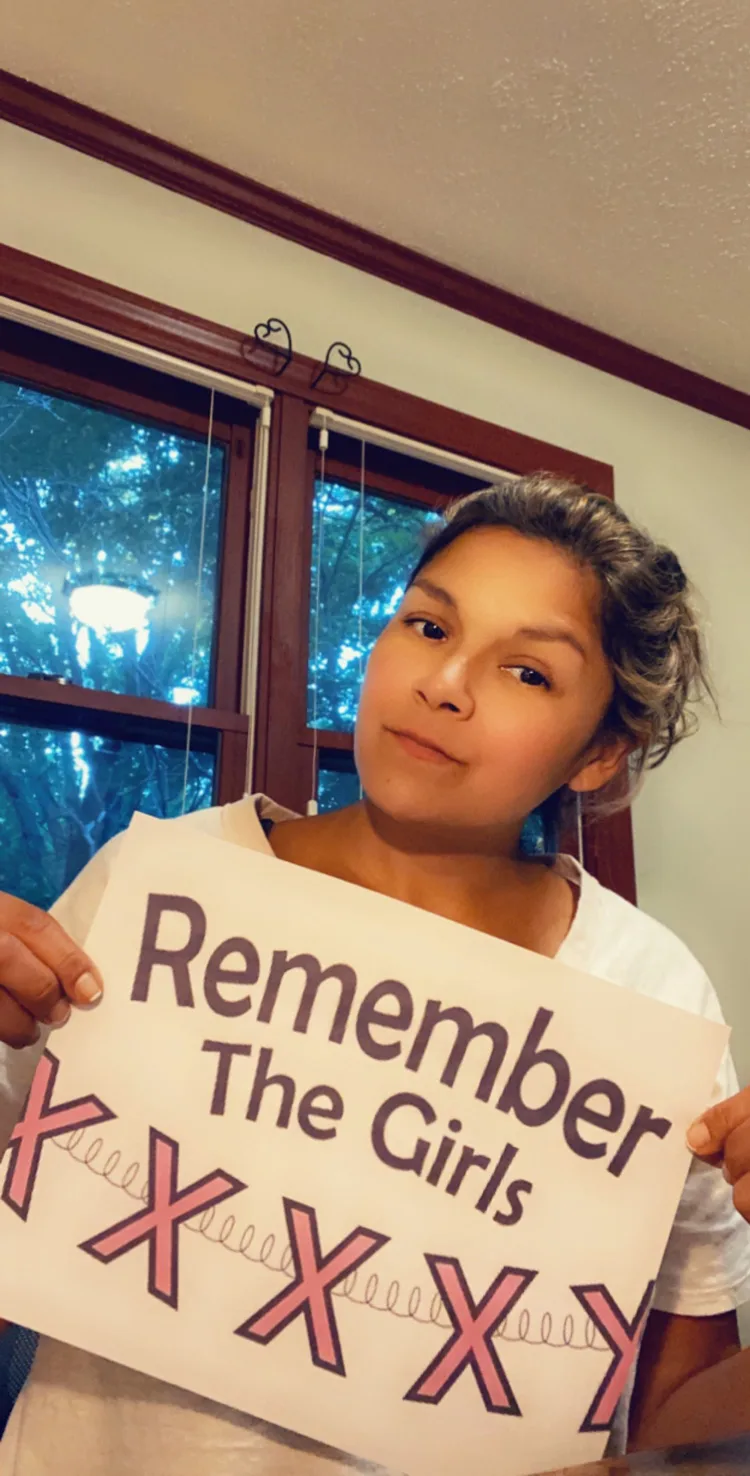 A woman in front of a window holding up a piece of paper with the Remember The Girls logo.