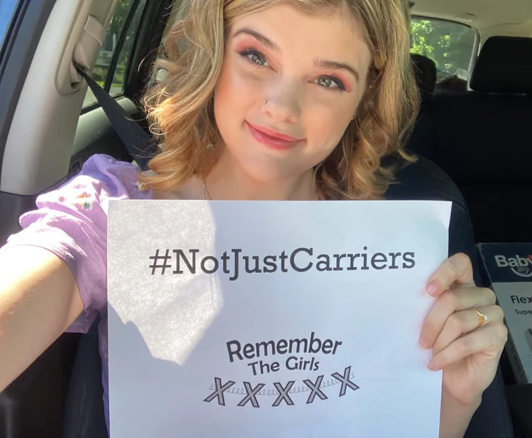 A community member sitting in the car holding up a sign that reads "#NotJustCarriers" above the Remember The Girls logo.
