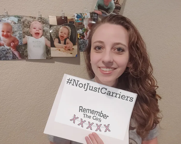 A woman in front of photos of her baby holding up a sign that reads "#NotJustCarriers" above the Remember The Girls logo.