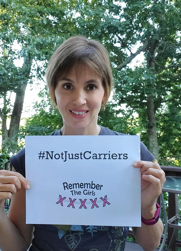A community member standing in front of trees holding up a sign that reads "#NotJustCarriers" above the Remember The Girls logo.