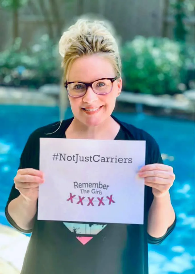A woman standing in front of a pool holding up a sign that reads "#NotJustCarriers" above the Remember The Girls logo.