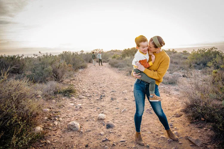 A woman holds her toddler son in her arms as they both laugh with the desert sun setting behind them.